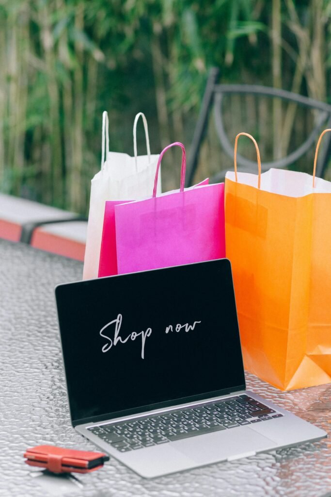 pexels-photo-6214155-6214155 Laptop displaying 'Shop now' with vibrant shopping bags on glass table outdoors.