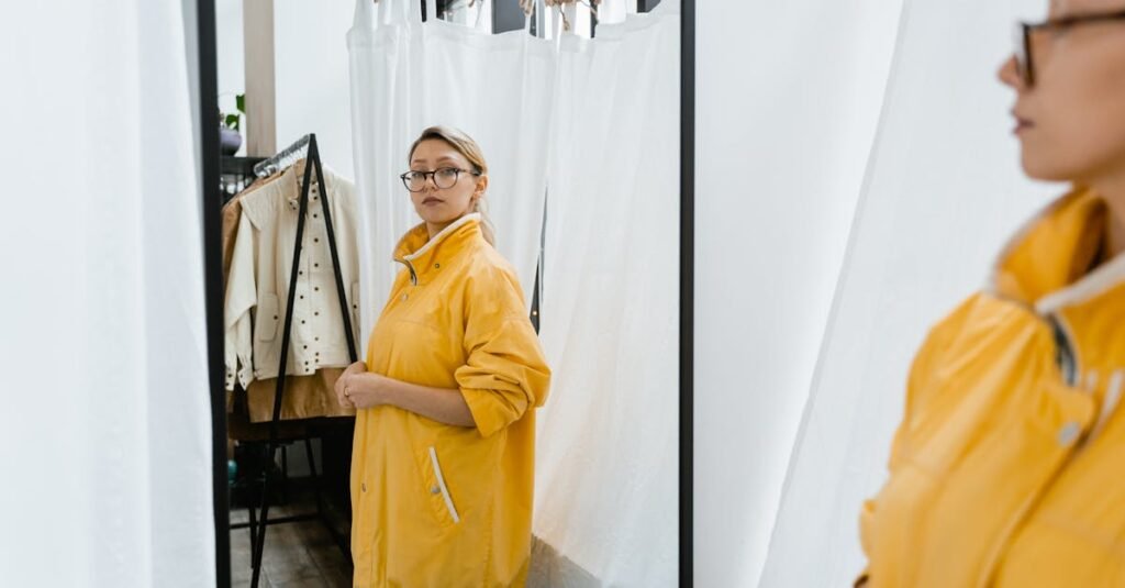 pexels-photo-7667439-7667439 A woman in a yellow coat examines her reflection in a fitting room mirror surrounded by trendy clothing.